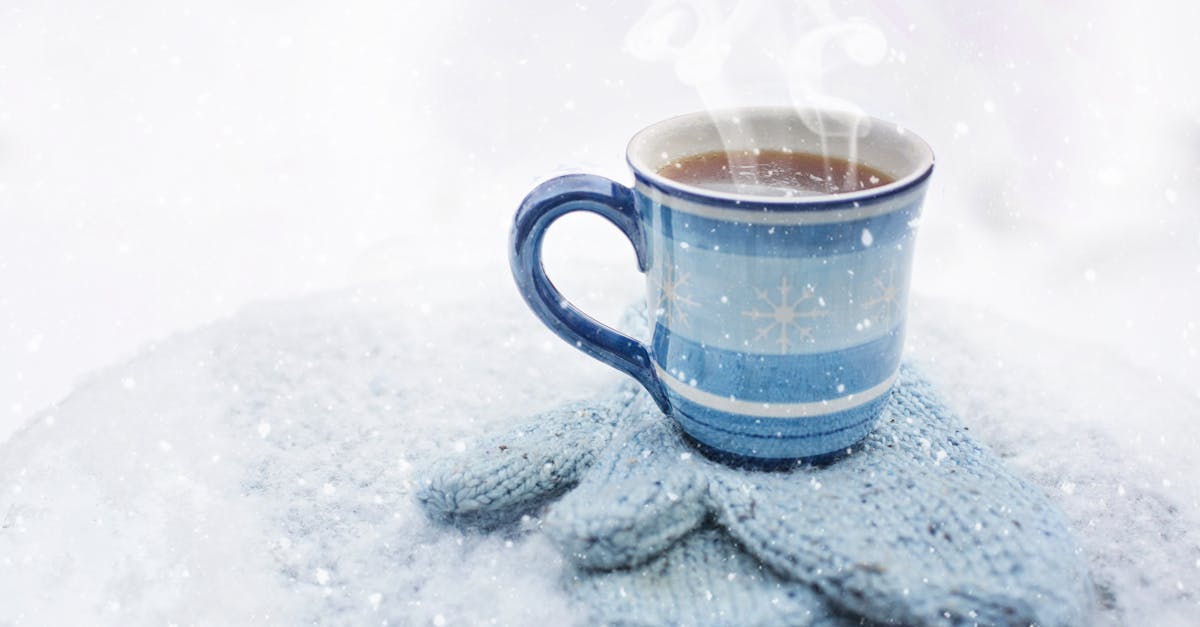 A steaming blue coffee mug sits on knitted gloves amid snowfall, evoking coziness.
