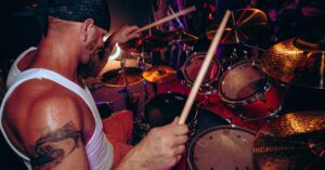 Bearded drummer energetically playing drums at a live music concert with tattoos and bandana.